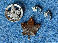 Photo showing two maple leaf brooches (one circular with cut-out leaf design and one solid maple leaf) alongside a pair of small maple leaf clip-on earrings on a blue fabric background.