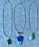 Three necklaces laid flat showing the pendants: green faceted glass teardrop on gold plated chain, blue triangular dichroic glass on silver chain, oval turquoise on silver chain.