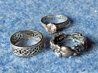 All three rings positioned on a blue textured surface: one with a large clear round gem, one with Celtic knotwork open design, and one with heart and floral decorative elements.