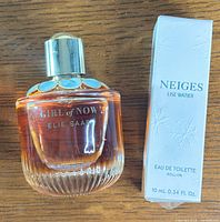 Bottle of Elie Saab Girl of Now perfume next to the boxed Lise Watier Neiges eau de toilette roll-on on a wooden surface.