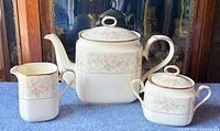 Full set of Noritake Willowbrook teapot, creamer, and lidded sugar bowl on blue cloth background showing floral pattern and gold trim.