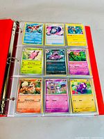 Open red binder displaying nine Pokemon cards per page in plastic sleeves, showing variety of Pokemon and Trainer cards with colorful artwork.