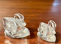 Both Swarovski crystal swan figurines placed side by side on wood surface, showing size difference and clear faceted crystal body