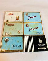 Top view of six vintage cigarette tins laid out in two rows of three, showing brands Sweet Caporal, Player's, Winchester, Black Cat, and Marcovitch with worn and aged surfaces.