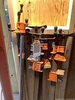 Full lot of seven 4-foot metal bar clamps with orange clamping heads and handles, standing against a wooden wall.