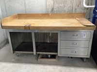Front view of the workbench showing entire butcher block top, stainless steel base, open compartments, and three metal drawers on the right side.