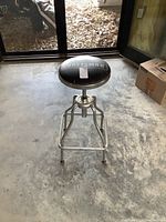 Front and side view of the Craftsman hydraulic adjustable shop stool placed on a concrete floor near glass doors showing full stool details including padded seat and metal frame.