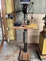 Front view of Delta ShopMaster DP400 drill press on metal stand with wooden work surface, lamp mount and accessories