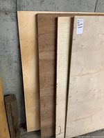 Four stacked plywood sheets leaning against a concrete wall, showing veneer faces including maple and oak.