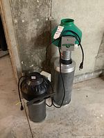 Two inline duct fans of different brands and designs on a concrete floor, one green topped HydroFarm model and a black topped Ivosun model with cords visible.