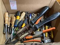 Box of assorted hand tools including screwdrivers with wooden and plastic handles, pliers, and cutting tools.