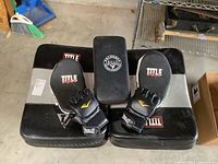 Photo showing two Title brand black and silver large kick pads, one smaller black LDU Company pad, and a pair of Everlast fingerless gloves.