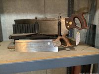 Side view of two hand saws with wooden handles and a metal mitre guide on a shelf