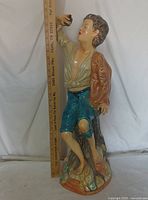 Full view of ceramic statue with measuring stick for scale showing approximately 28 inches tall, displaying the full figure of the boy in colored clothing holding a shell.