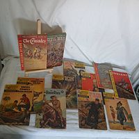 Full spread of 12 vintage comic and illustrated books with covers visible, arranged on cloth background with a wooden display stand showing two books upright.