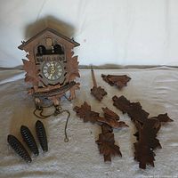 Front view of coo-coo clock with detached carved wooden decorative parts and pine cone weights laid out on surface.