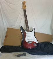 Full view of red sunburst electric guitar standing with black carrying case and shipping box in background