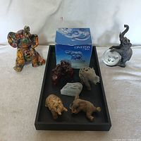 Overview of all seven elephant figurines displayed in a black tray including one in original packaging and six loose figurines