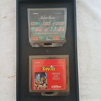 Two game cartridges: Tandy Joko-Ban (top) and Activision Rampage Smash and Trash (bottom) in clear plastic cases on a black background.