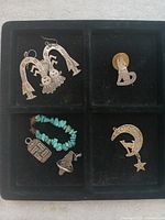 Top-down photo showing four jewelry pieces in a black divided tray including silver earrings with winged figure, a small pendant shaped like a coyote, a crescent moon pendant with a star, and a turquoise chip bracelet with two additional pendants.