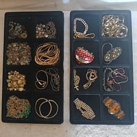 Two black trays with multiple compartments filled with assorted costume jewelry including bracelets, necklaces, earrings, and pendants in various styles and materials.