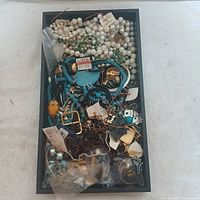Top view of box containing assorted jewelry including pearl and turquoise necklaces, gold and silver tone earrings, and mixed craft materials.