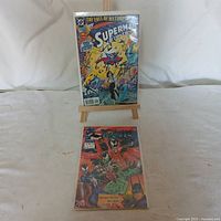 Photo of both signed comic books, 1994 Superman 'The Fall of Metropolis' displayed upright on an easel and 1992 Yawie 'Splatman Returns' lying flat on table.