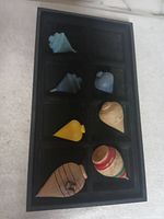 Seven wooden spinning tops in a black divided display tray with various colors and shapes