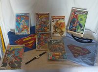 Full lot showing seven Superman comic books arranged around two t-shirts (blue and red) and a black Superman wristwatch on a flat surface
