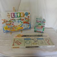 Three unopened board games displayed: The Game of Life on an easel, Monopoly Golf Edition flat, and Monopoly Diced upright.