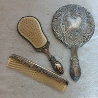 Set of three vintage hair grooming items: a comb, a brush, and a hand mirror, all with ornate silver-tone metal decorative handles.