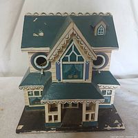 Front view of the vintage birdhouse showing house shape, porch, scalloped roof trim, two round bird entry holes with perches, painted in blue, cream, purple colors with paint chipping.