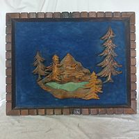 Front view of the wooden artwork showing carved trees, mountain, and pond on blue felt background framed in wood