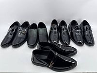 Seven pairs of black patent leather slip-on dress shoes arranged in two rows, showing various styles including metal buckle accents and smooth upper finish.