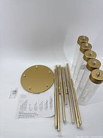 All components of the gold candle stand set including circular base, acrylic tubes, metal rods, screws, and instructions laid out on white background.