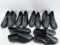 Five pairs of black leather loafers arranged in two rows on white surface. Each pair looks new with a minimalist design and silver emblem on the upper.