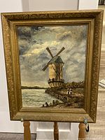 Full view of the framed oil painting showing a windmill on a body of water with a cloudy sky and two figures on the dock.