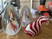 Photo shows two clear teardrop-shaped art glass paperweights and one red and white striped small glass sculpture arranged on a surface.
