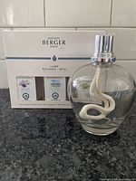 Clear glass round Lampe Berger fragrance lamp with visible wick inside, placed before the refill pack box with two refill bottles shown: Lavender and Linen scented refills.