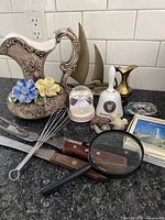 Full lot arranged on kitchen counter including ceramic pitcher, brass sailboats, brass vase, bell, kitchen utensils, magnifying glass, souvenirs, and small carved figures.