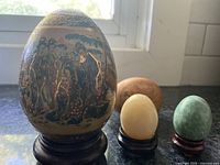 Three vintage decorative eggs: one large painted egg with Asian-style artwork, cream-colored egg and green egg on wooden stands.