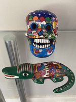Photo of colorful hand-painted skull and green alligator figurines with cityscape and floral patterns on glass shelf.