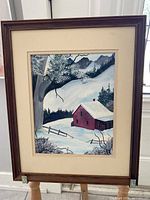 Full front view of a framed painting showing a snow-covered landscape with a red house, a large tree, fence, and mountain backdrop.