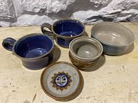 Five signed studio pottery pieces displayed on a wooden table with a stone wall background showing two blue glazed cups, a small pot, a bowl and a lid with a sun face design.