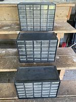 Three plastic storage boxes with multiple small drawers arranged on wooden shelves for photo. Boxes have transparent drawers with black framing.