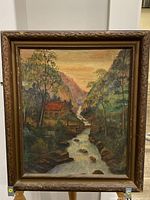 Framed oil painting showing entire artwork with mountain scenery, cabin, and water under warm sky.