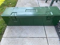 Full view of the elongated green metal box placed on stone pavement, showing overall shape and military marking.