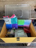 Wooden bin with transparent lid, filled with brightly colored plastic laundry clips, white hooks, and other accessories.