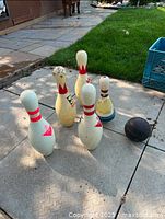 Five vintage bowling pins arranged standing on a paved surface with one bowling ball nearby.