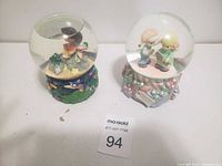 Two globes side by side on a flat surface: one Dora the Explorer music globe with colorful nature base, the second with two children on a floral pastel base.